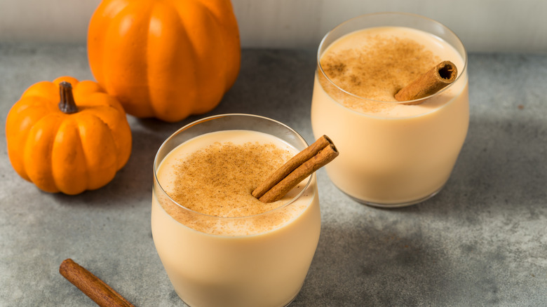 Two glasses of pumpkin spice eggnog next to two pumpkins