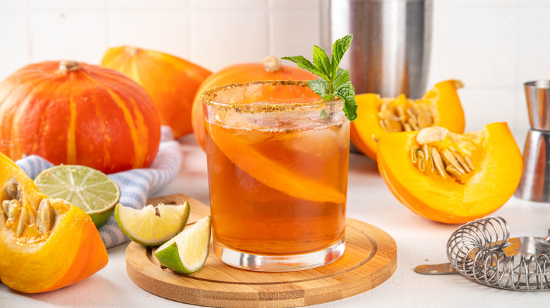A pumpkin margarita in a tumbler surrounded by pumpkin pieces and lime wedges