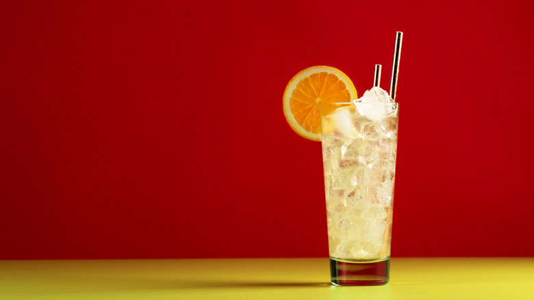 A glass of Tom Collins with an orange wedge garnish against a red background