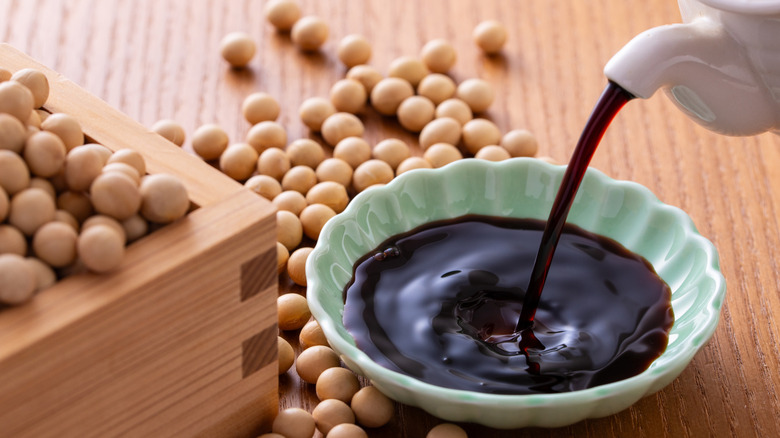 Soy sauce being poured in a dipping bowl surrounded by whole soy beans