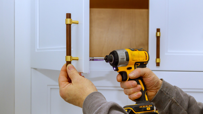 A pair of hands using a power drill to attach a handle to a white cabinet door