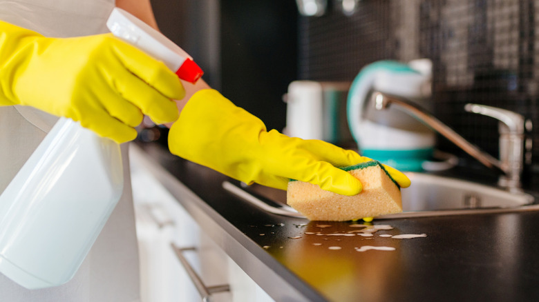 A woman using a spray bottle and orange sponge to clean a kitchen
