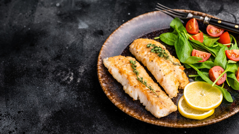 Roasted cod fillet portions shown on a dark brown plate against a mottled black countertop garnished with lemon slices with a simple salad of baby spinach and quartered cherry tomatoes