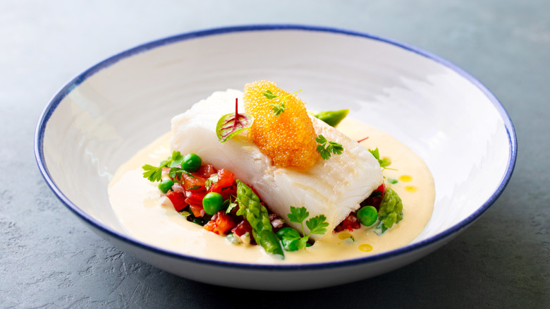 An elegant meal of halibut fillet on a bed of baby vegetables and cream sauce topped with a quenelle of pike roe served in a simple white bowl with blue rim