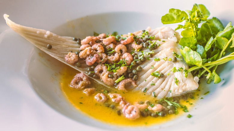 Skate wing as served in an upscale restaurant in butter sauce and garnished with small shrimp capers chopped herbs and a salad of baby greens