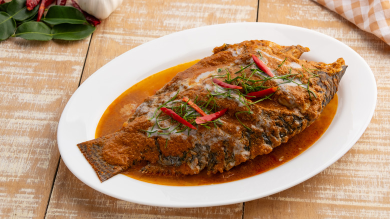 Whole grilled tilapia on a simple white plate in Thai-style red curry sauce garnished with herbs and slivers of hot red pepper