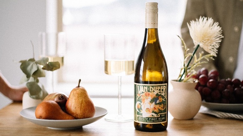 bottle of white Van Duzer Pinot Blanc wine on table with fruit, flowers, and wine glasses
