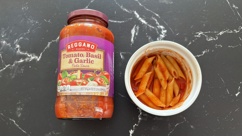Aldi tomato, basil, and garlic sauce jar next to pasta in bowl on a dark countertop