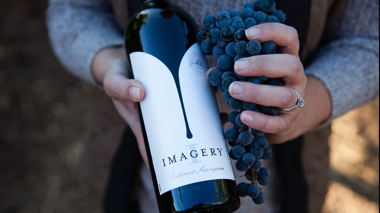 Hands holding a bottle of Imagery cabernet sauvignon and cabernet grapes.