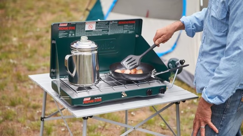 Person cooking on Coleman 120-square-inch green portable liquid propane grill outdoors in front of a tent
