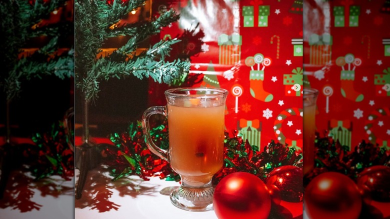 Orange-red cocktail in a toddy glass with a bright red ornament in front of it and a Christmas tree and wrapping paper behind