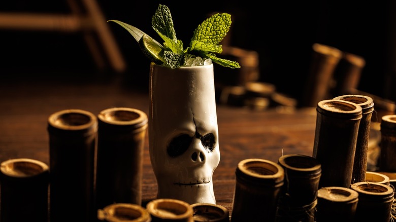 Tall skull-shaped tiki glass overflowing with crushed ice and stuffed with mint leaves and lime, surrounded by about a dozen tall, thin shot glasses