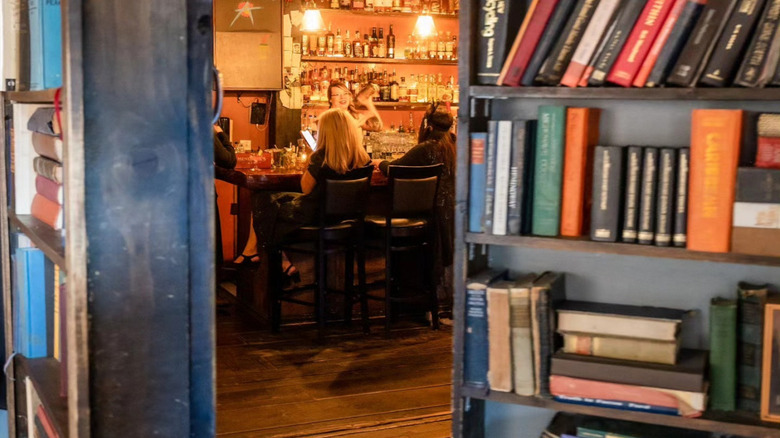 Bookshelf concealing hidden bar lined with patrons sipping cocktails sitting in front of a bartender using a cocktail shaker