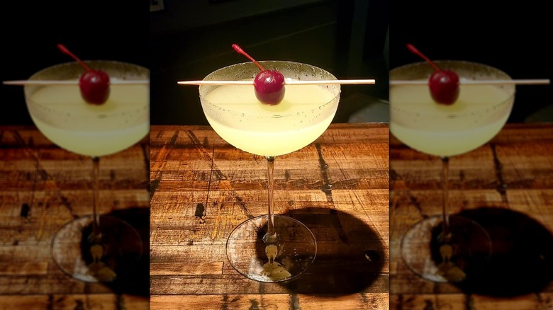 Lemon-yellow cocktail in a delicate coupe glass, with a cherry garnish balanced on a toothpick, sitting on old wooden bar