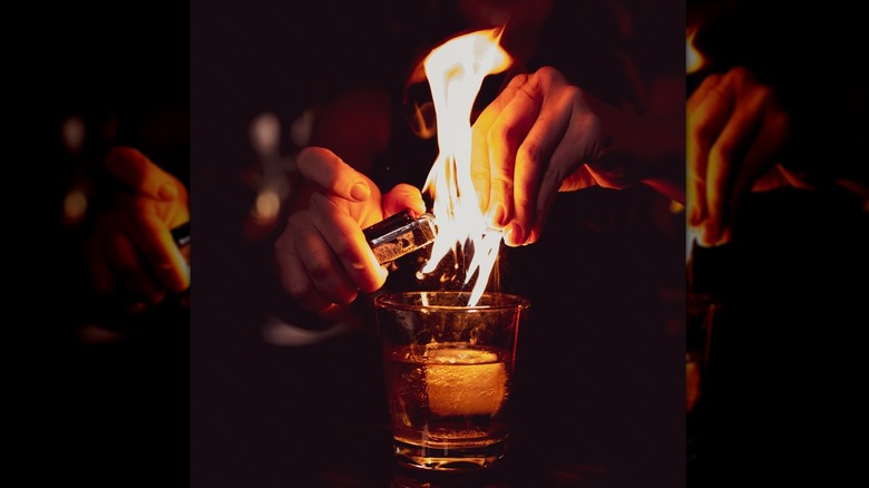 Hand holding a lighter setting a whiskey cocktail with a giant ice cube into fire, flames leaping out of the glass