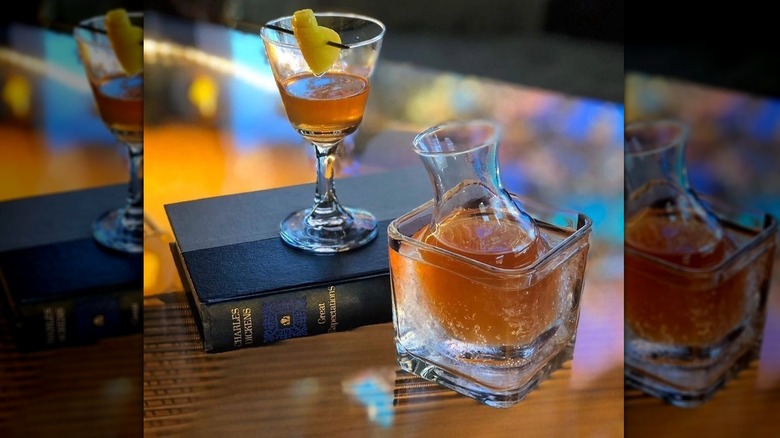 Half-empty cocktail with lemon garnish sitting atop a copy of Charles Dickens's "Great Expectations," next to a mini carafe filled with a cocktail in a glass of water