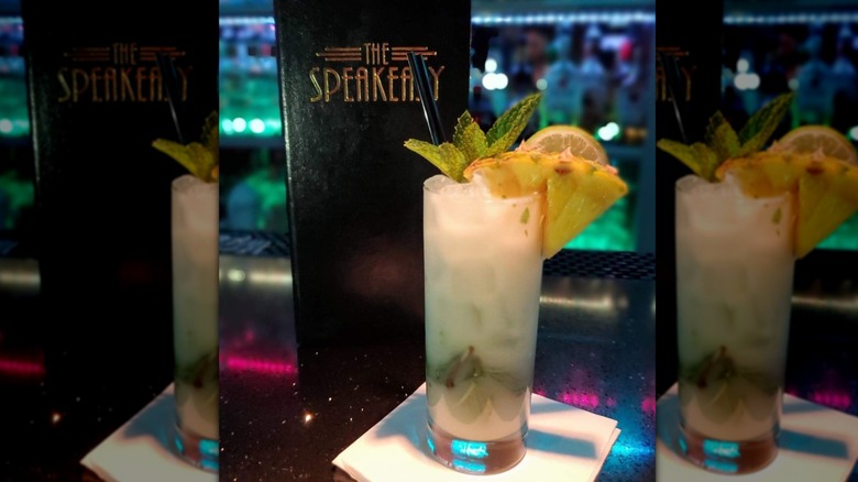 Frothy pineapple mojito in a tall rocks glass on white cocktail napkin in front of a slim black menu standing up that read "The Speakeasy"