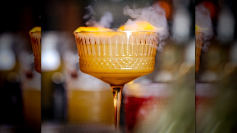 Bright orange cocktail in ornate glass with curls of smoke billowing from it