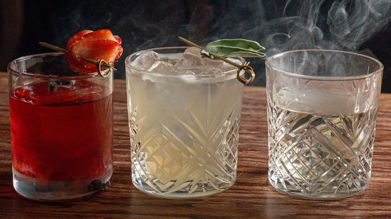 Line of three cocktails in rocks glasses; one red with strawberry garnish, one pale yellow with sage leaf garnish, and one clear liquid emitting curls of smoke