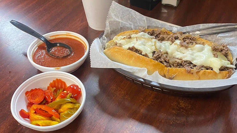 Cheesesteak in basket next to cup of marinara sauce and cup of grilled peppers and carrots all on wooden table