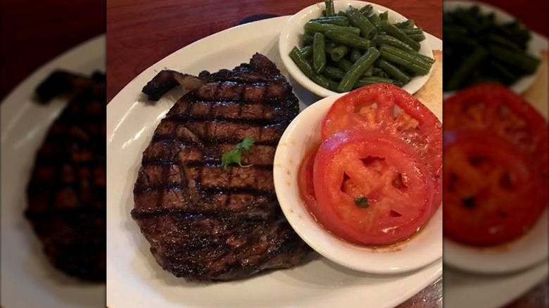 Tom's Choice marinated rib eye steak on white plate next to small white bowls of tomato slices and green beans