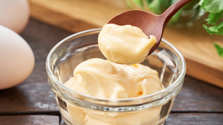 A wooden spoon filled with mayonnaise and a glass bowl filled with creamy mayonnaise