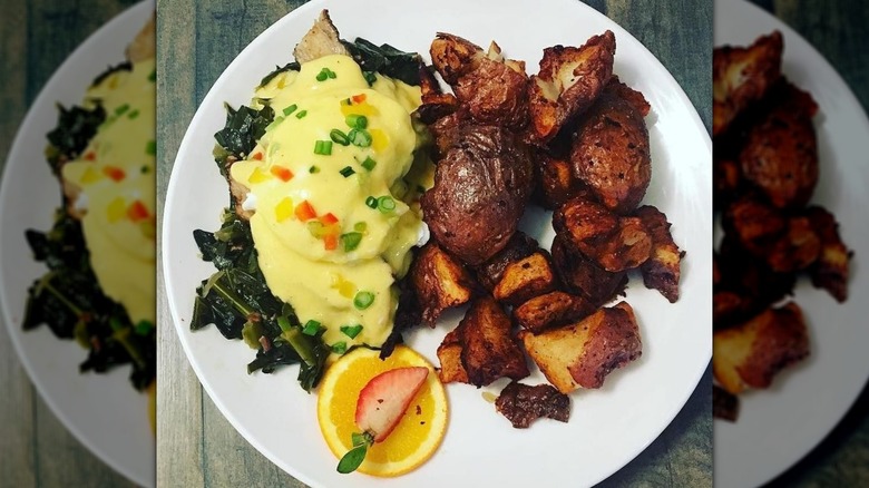 Bluegrass Benny eggs Benedict loaded with hollandaise sauce dripping over eggs on a pile of collard greens, a side of crispy potatoes, and a sliced orange on a white plate