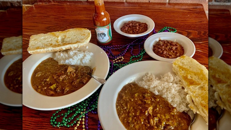 White bowls containing gumbo and red beans and rice, with a half slice of French bread perched on the side, a bottle of hot sauce, and Mardi Gras beads on a wooden table