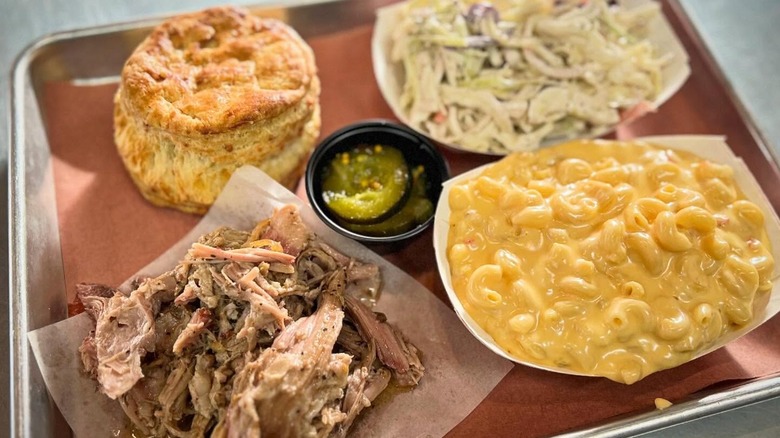 Cornbread muffin, coleslaw, pickles, mac and cheese, and pulled pork in separate containers on a metal serving tray