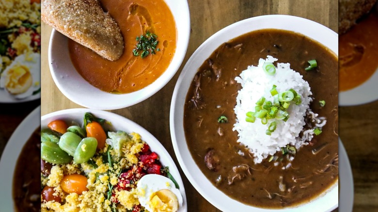 Three white bowls; one containing a pile of white rice sitting in dark brown gumbo, another with an orange/red soup and crusty bread, and the third with a salad containing hard-boiled egg, cherry tomatoes, and baby peppers