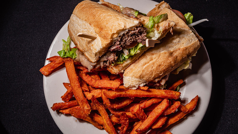 Two stacked halves of a juicy beef debris po'boy on French bread with shredded lettuce and a side of sweet potato fries on a white plate against a black background