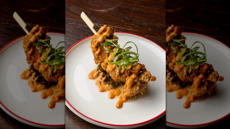 Juicy chicken on a wooden skewer on a white plate, dripping with orange-brown sauce and topped with curls of green onion