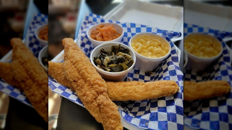 Basket lined with a blue gingham paper containing long pieces of breaded and fried whiting fish and 3 round containers of collard greens, mac and cheese, and yams