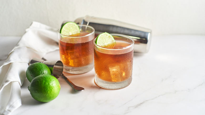 Two rocks glasses of cola with shaker and whole limes.