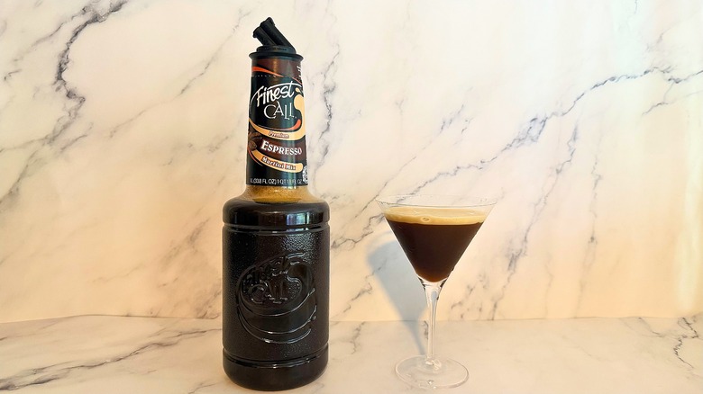 Bottle of Finest Call espresso martini mix next to espresso martini in glass on marble surface