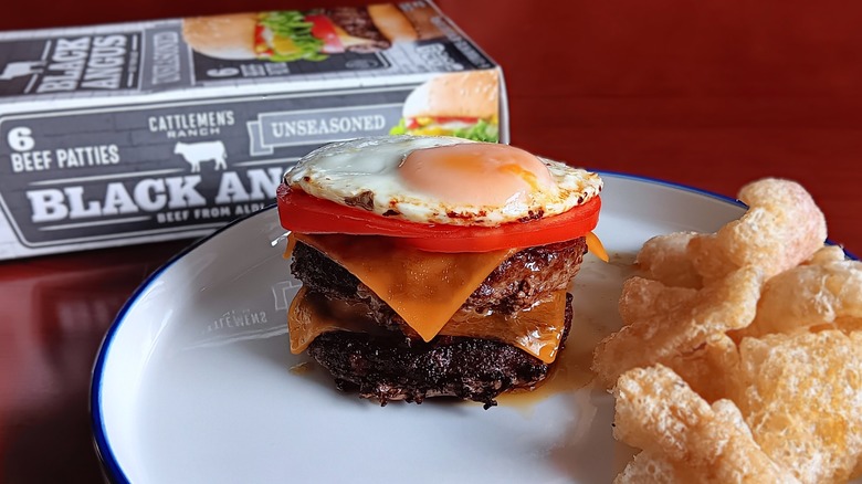 double cheeseburger topped with tomatoes and sunny side up egg with a side of pork rinds on white plate in front of Cattlemen's Ranch frozen beef patties box