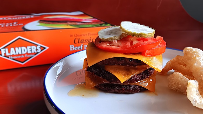 Triple cheeseburger with tomatoes and pickles on a white plate with blue trim in front of a Flanders frozen beef patties box