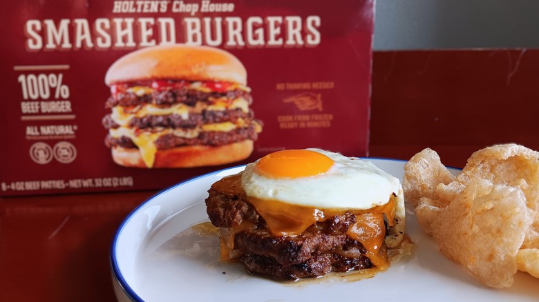 triple cheeseburger topped with sunny side up egg with a side of pork rinds on white plate in front of Holten's Chop House frozen smashed burgers box