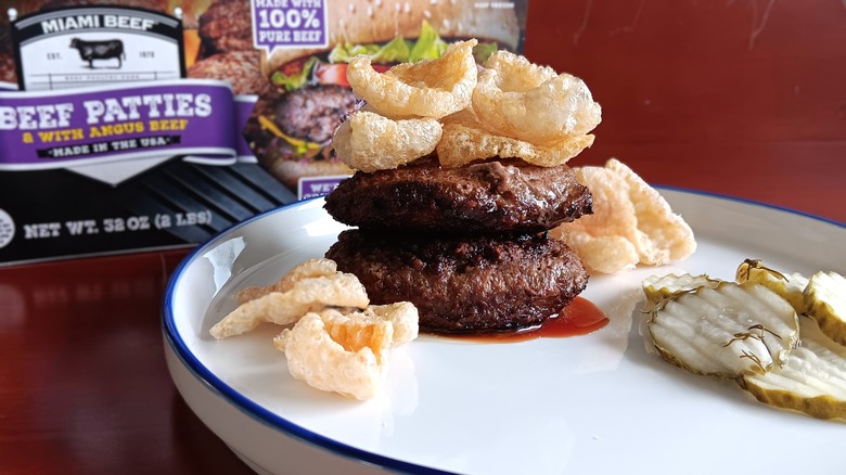 double burger topped with pork rinds with a side of pickles on white plate in front of Miami Beef frozen beef patties box