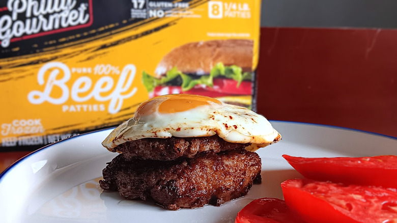 double burger with sunny side up egg on top with side of tomatoes on white plate in front of Philly Gourmet beef burgers box