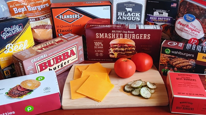 various brands of frozen beef burgers surrounding a cutting board with slices of cheese, a couple of tomatoes, and pickles