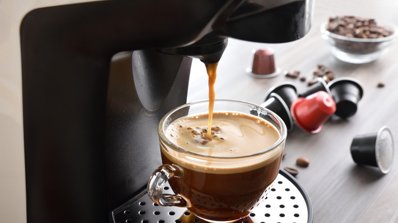 Machine pouring coffee into clear cup with coffee pods and beans in background