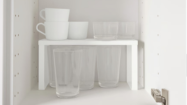 A white shelf insert from Ikea in a cupboard storing mugs and short glasses on top and tall glasses below