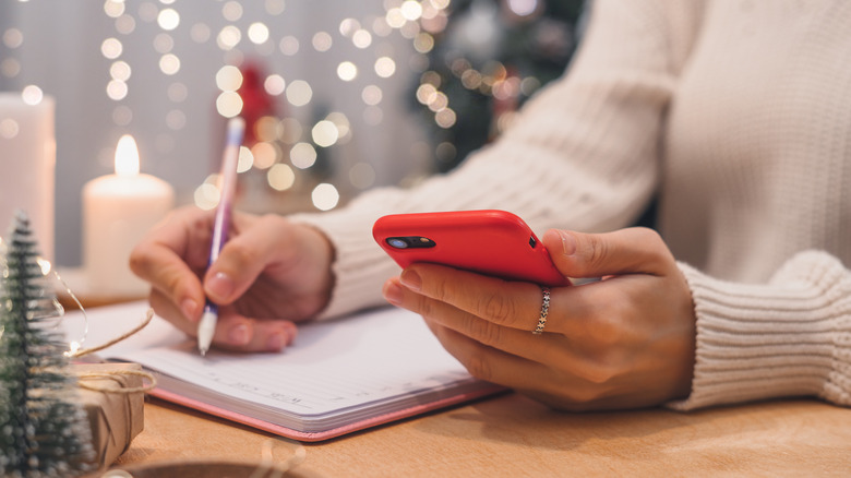 Woman writing notes while holding her phone, with Christmas decorations in the background