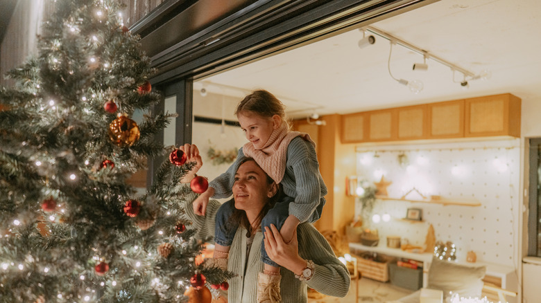 Woman decorating a Christmas tree with a child on her shoulders