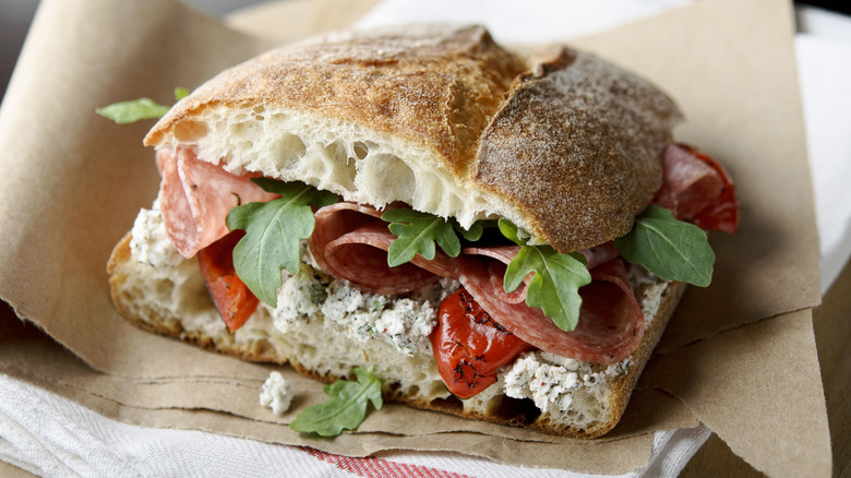 A salami, tomato, and greens sandwich on a brown wrapper.