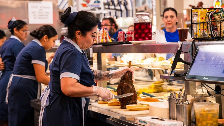 Woman in uniform with arm tattoos cutting meat on bone for a sandwich.