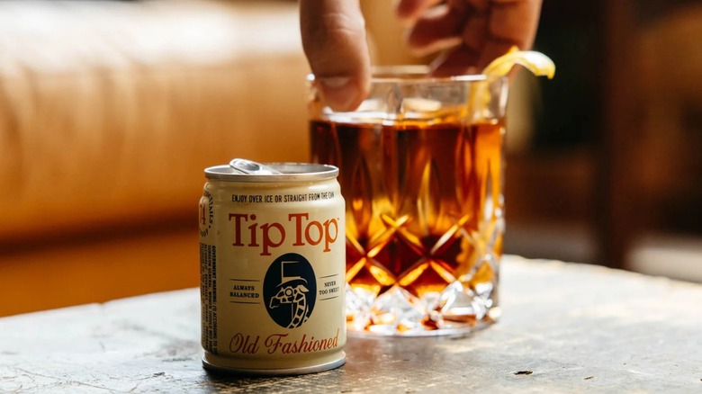 A can of Tip Top Old Fashioned next to a poured glass with a hand reaching down to lift it.