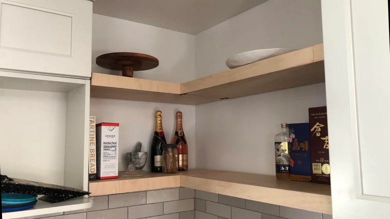 Two tiered plywood shelf holding alcohol bottles mounted on wall between white cabinets