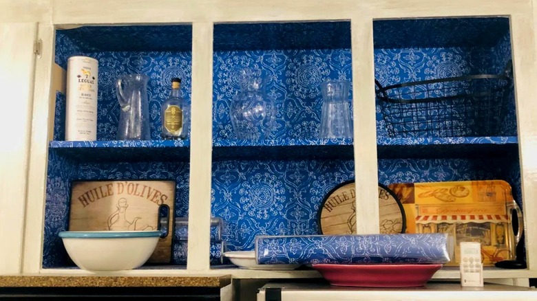 Wall shelves with blue patterned wallpaper backing holding glass vases and decorative signs in kitchen